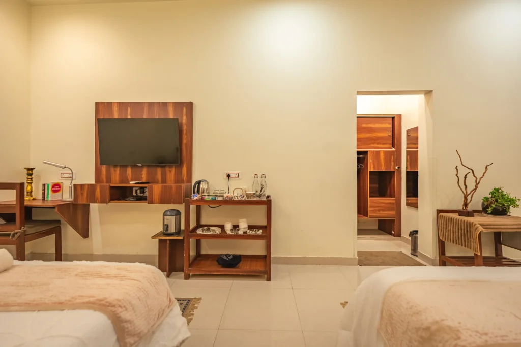 Well-appointed double deluxe room at Pristine Woods, a Luxury Resort in Dehradun, featuring twin comfortable beds, a modern wall-mounted TV, elegant wooden furniture, in-room tea and coffee setup, spacious wardrobe area and a calm, premium ambience.