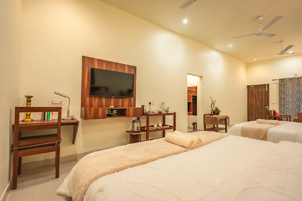 Spacious luxury room interior at Pristine Woods, a Luxury Resort in Dehradun, featuring a plush king-size bed, modern wall-mounted TV, elegant wooden furnishings, in-room amenities and a serene ambience for a premium stay.
